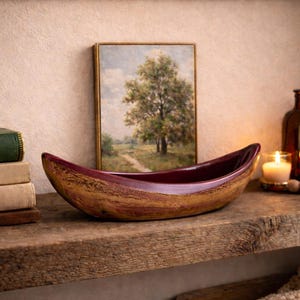 Op de afbeelding: Een decoratieve houten kom met een bordeauxrode binnenkant staat op een rustieke houten plank. Een ingelijste landschapsschilderij, een kaars en een donkere glazen fles staan ook op de plank. Links staat een stapel boeken.