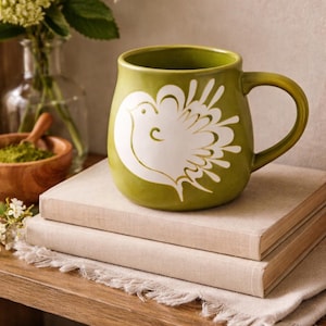 May include: Green ceramic mug with a white bird design. The mug has a rounded shape and a large handle. It sits on top of two books with beige covers. The mug is part of a home decor collection.
