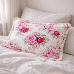 May include: Rectangular pillow with a floral pattern of pink and red roses on a white background. The pillow has a quilted border and is placed on a white bed. The pillow is a decorative bedding item.