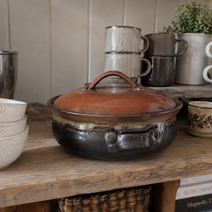 May include: A round, lidded ceramic casserole dish featuring a glossy, deep-brown base with two side handles and a contrasting reddish-orange lid. The pot is centred among various speckled ceramic bowls and mugs on a wooden shelf.