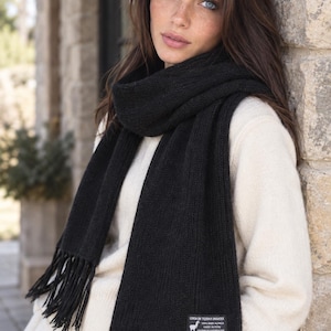 May include: A black knit scarf featuring a ribbed texture and fringe detailing at both ends. A rectangular label is stitched onto the lower corner, displaying the text Linea de Tejidos Inkatex, 100% Baby Alpaca, Made in Peru, Calidad de Exportacion.
