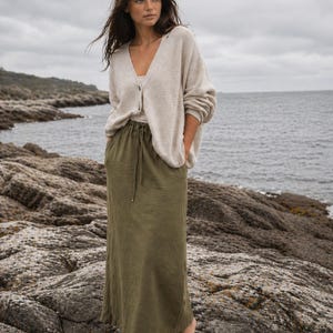 May include: A woman in a beige cardigan and olive green maxi skirt stands on rocks by the sea. The cardigan has a V-neck and buttons. The skirt is a solid colour with a drawstring waist. The background shows the ocean and a cloudy sky.