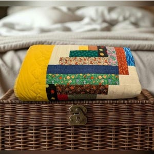 May include: A folded quilt with a patchwork design, featuring a yellow quilted edge and a central section with various fabric strips in blue, green, floral, and patterned designs. The quilt is resting on a brown wicker chest.