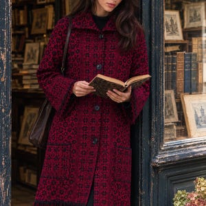 May include: A long-sleeved, knee-length coat in a deep red and black patterned fabric. The coat features a repeating geometric floral design, a wide collar, four dark buttons down the front, two large patch pockets, and a fringed hemline.