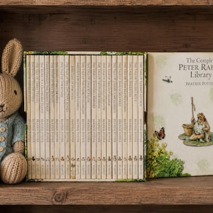 Op de afbeelding: Een gebreide konijnenpop met een blauw jasje zit naast een boxset van Beatrix Potter-boeken. De boekenreeks heet "The Complete Peter Rabbit Library" en bevat illustraties van Peter Rabbit.