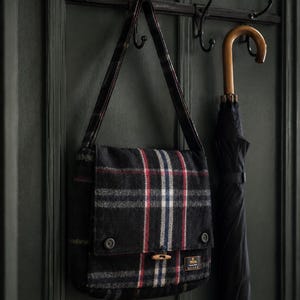 Peut inclure: Un sac messager à carreaux noirs avec des rayures rouges, blanches et bleues est suspendu à une porte vert foncé. Le sac a une fermeture à bascule en bois et une bandoulière. Un parapluie noir est également accroché.