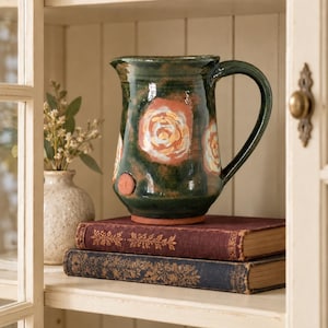 May include: A dark green ceramic pitcher featuring hand-painted floral motifs in shades of orange, yellow, and white. The pitcher has a curved handle, a flared rim, and a small circular clay stamp on the lower side, resting atop two stacked books.