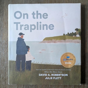 May include: Book cover for "On the Trapline" featuring an illustration of a person and child gazing at a seascape. The cover displays the title, author names "DAVID A. ROBERTSON" and "JULIE FLETT", and a "ggbooks Winner" seal.