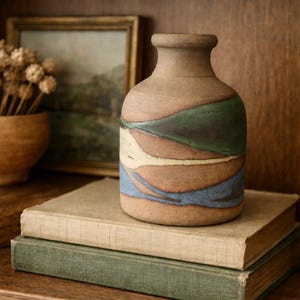 May include: A handcrafted ceramic vase with a brown base and colourful bands of green, blue, and cream. The vase sits on a stack of two vintage books with a framed landscape painting in the background. The vase is approximately 15 cm tall.