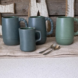 May include: Four ceramic mugs in shades of blue and green are displayed on a beige linen cloth. Two small silver spoons are placed in front of the mugs. The mugs have a textured finish and unique handle designs. The background features wooden letters.