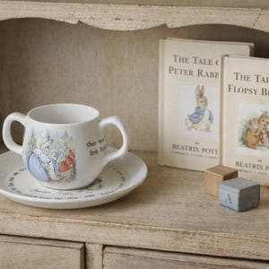Puede incluir: Un juego de taza y platillo de cerámica blanca con una ilustración de Peter Rabbit. Dos libros de Beatrix Potter, bloques de madera y una figura de conejo de madera en un estante.