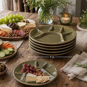 May include: Six olive green divided serving plates, each 23 cm in diameter, are stacked on a wooden table. The plates have a unique design with four sections. A charcuterie board and other food items are also on the table.