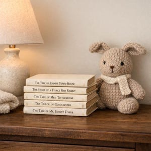 Könnte beinhalten: Ein Stapel von fünf alten Büchern mit Titeln wie "The Tale of Johnny Town-Mouse" und ein gestrickter Hase mit Schal. Die Bücher sind beige mit goldener Schrift. Der Hase ist hellbraun.