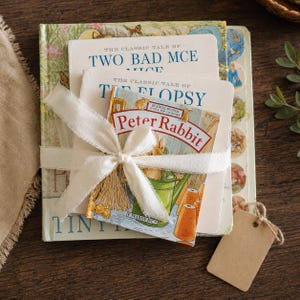 Könnte beinhalten: Ein Stapel Kinderbücher, darunter "Peter Rabbit", mit einem cremefarbenen Band zusammengebunden. Die Bücher haben farbenfrohe Illustrationen und Titel wie "Zwei böse Mäuse" und "Die Flopsy-Hasen". Ein leeres Etikett liegt daneben.