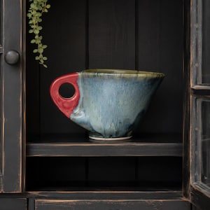 Puede incluir: Una taza de cerámica grande, hecha a mano, con un esmalte azul y verde. La taza tiene un asa roja y se encuentra en un estante negro dentro de un armario oscuro. Una ramita de vegetación cuelga encima de la taza.