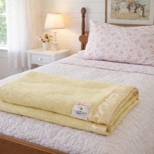 May include: A folded yellow blanket with a satin border rests on a bed with a white quilted bedspread and a floral patterned pillow. A framed picture and a lamp are visible in the background.