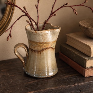 May include: A ceramic pitcher with a flared rim and a curved handle, featuring a speckled cream-colored upper section and a smooth, earthy tan glaze on the lower body. The pitcher contains several thin, dark red branches with small buds.