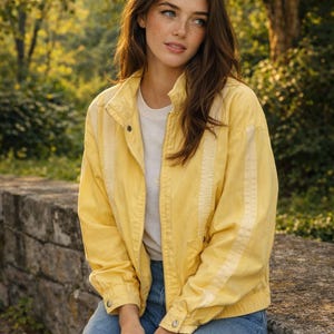 May include: A yellow jacket with vertical stripes on the sleeves and a white t-shirt. The jacket has a zip closure and buttoned cuffs. The person is wearing blue jeans. The background is a blurred view of trees and foliage.