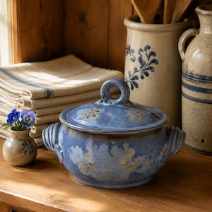May include: A blue ceramic lidded casserole dish with a mottled finish and floral accents, featuring two side handles and a circular lid knob. The pot displays a textured glaze with light blue and tan patterns, sitting alongside a small bud vase.