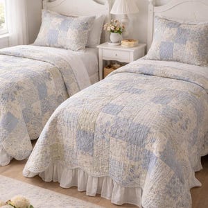 以下が含まれることがあります： 白いヘッドボードとマッチした寝具を備えた2つのシングルベッド。キルトと枕カバーは、青、クリーム、ペールグリーンの色合いのパッチワークデザインが特徴です。白いフリル付きベッドスカートがエレガントさを添えています。