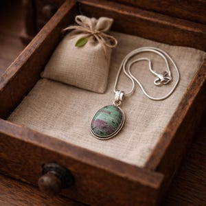 May include: A silver necklace with a large oval pendant featuring a green and red stone, displayed in a wooden drawer with a small burlap bag. The drawer is lined with beige fabric.