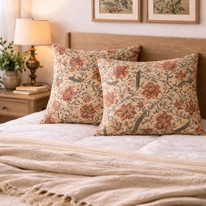 May include: Two square cushions with an orange, red, and blue floral pattern on a cream background. They sit on a white bed with a beige fringed throw. Two framed floral prints hang above the bed, creating a cosy bedroom scene.