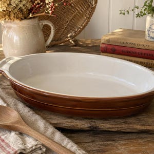May include: An oval ceramic baking dish with a glossy white interior and a rich brown exterior. The dish features integrated handles on both ends and a smooth, glazed finish. It is a functional kitchen vessel designed for oven use and serving.