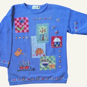 Op de afbeelding: Een lichtblauwe trui met geborduurde patches met tuin- en natuurtaferelen. De patches bevatten een vogel, een boom, een huis, een schildpad en bloemen. De trui heeft lange mouwen en een ronde hals, met decoratief borduurwerk op de manchetten en zoom.