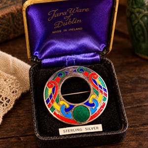 May include: A circular sterling silver brooch featuring colorful enamel inlays of red, blue, green, and yellow, with a central green stone. The brooch is housed in a black presentation box labeled "Tara Ware of Dublin" and "STERLING SILVER."