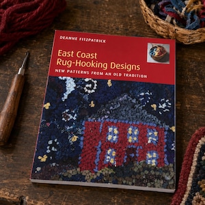 May include: A softcover book titled East Coast Rug-Hooking Designs by Deanne Fitzpatrick. The cover features a dark blue night scene with a red house and yellow windows. A rug hook with a wooden handle sits to the left of the book.