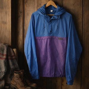 May include: A blue and purple anorak jacket with a hood, hanging on a wooden hanger. The jacket has a color block design, with a blue upper section and a purple lower section. The jacket is made of a lightweight material.