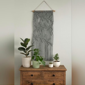 May include: A grey macramé wall hanging with a leaf design hangs from a wooden dowel. The wall hanging is above a wooden chest of drawers with several potted plants. The plants include a large-leafed plant in a white pot, a green potted plant, and a small succulent.