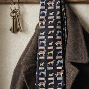 May include: A navy blue tie featuring a repeating pattern of various dog breeds, including terriers and greyhounds. The tie is hanging on a wooden coat rack next to a brown coat. The keys are hanging on the left side of the image.