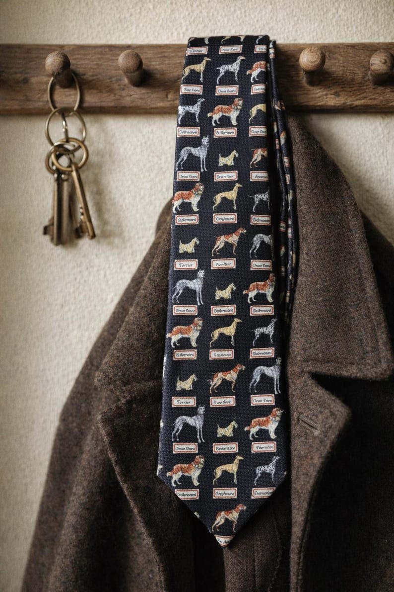 May include: A navy blue tie featuring a repeating pattern of various dog breeds, including terriers and greyhounds. The tie is hanging on a wooden coat rack next to a brown coat. The keys are hanging on the left side of the image.