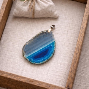 May include: A blue agate pendant with a silver bail sits on a linen surface in a wooden tray. A small beige pouch tied with twine and a green leaf is in the background. The agate features shades of blue and white.