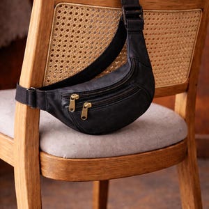 May include: A black leather bum bag with two zipped compartments and gold-coloured hardware hangs on a wooden chair. The chair has a woven cane back and a grey upholstered seat. The bum bag's strap is black.