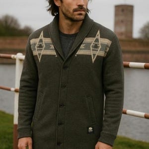 May include: Dark green button-up coat with a shawl collar and geometric pattern across the shoulders. The coat has a small logo patch and is worn over a gray shirt. The coat is knee-length and has two front pockets.