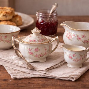 May include: A porcelain tea set with pink rose detailing, comprising a sugar bowl, creamer, and serving jug. The set is displayed on a linen cloth, with scones and jam in the background. The set is white with gold trim.