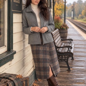May include: A grey zip-up jacket with a collar and two front pockets, worn over a cream-colored turtleneck sweater. The outfit includes a brown and grey plaid midi-length wrap skirt and knee-high brown leather boots with a block heel.