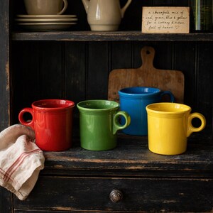 May include: Four colorful ceramic mugs in red, green, blue, and yellow are displayed on a dark wooden shelf. The mugs have rounded handles and a classic design. A small card with handwritten text is visible. A red and white striped cloth is on the left.