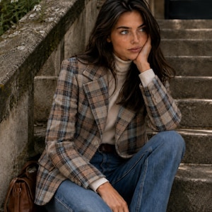 Op de afbeelding: Een geruite blazer in tinten beige, bruin en blauw, gedragen over een crèmekleurige coltrui en een blauwe spijkerbroek, vergezeld van een bruine leren handtas. De blazer heeft een ingekeepte revers, lange mouwen met knopen en een enkele knoopsluiting.