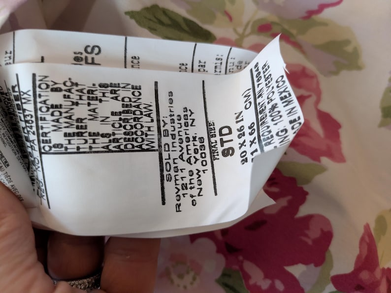 May include: A white tag with black text, including the words "CERTIFICATION" and "FINAL SIZE STD 20 X 26 IN". The tag is partially covering a floral fabric with pink and green flowers.