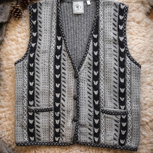 Op de afbeelding: Een grijze gebreide vest met een V-hals en knoopsluiting. Het vest heeft een geometrisch patroon in zwart en wit, met zwarte randen en zakken. Het wordt gepresenteerd op een getextureerd, lichtgekleurd oppervlak.