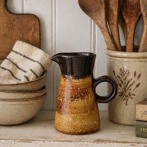 May include: A handcrafted ceramic jug with a dark brown top and a speckled, textured body in shades of brown and yellow. The jug has a curved handle and a spout. Other items include wooden spoons, bowls, and a decorative crock.