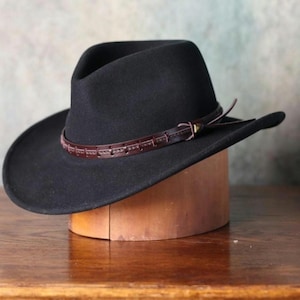 Pode incluir: Um chapéu de cowboy preto com uma faixa e fivela de couro castanho. O chapéu é exibido em um bloco de chapéu de madeira, mostrando sua forma. Possui uma aba larga e um estilo western clássico.