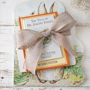 Könnte beinhalten: Ein Geschenkset mit einem Kinderbuch mit dem Titel "The Tale of Mr. Jeremy Fisher" von Beatrix Potter, gebunden mit einem Jutegarnband. Das Buch ist in einer dekorativen Karte mit einer Kaninchenillustration und einer Biene eingebettet.