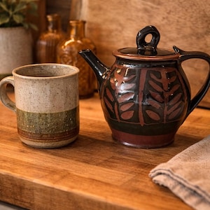 Peut inclure: Un ensemble tasse et théière en céramique. La tasse a un dessus beige moucheté, une bande centrale verte et une bande inférieure marron. La théière est noire avec un couvercle marron et un motif de feuilles rouge et marron.