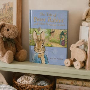 Könnte beinhalten: Ein gebundenes Buch mit dem Titel The Tale of Peter Rabbit and other Favourite Stories von Beatrix Potter, mit blauem Einband und einer Illustration von Peter Rabbit, neben einem braunen Plüschhasen und einem Holzhasen auf Rädern.