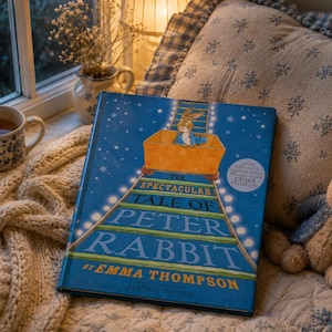 Könnte beinhalten: Ein blaues Buch mit dem Titel "The Spectacular Tale of Peter Rabbit" von Emma Thompson, mit einer Kaninchenillustration auf dem Cover. Ein Stoffkaninchen, eine Tasse Tee und eine cremefarbene Strickdecke sind ebenfalls zu sehen. Gemütliche Szene.