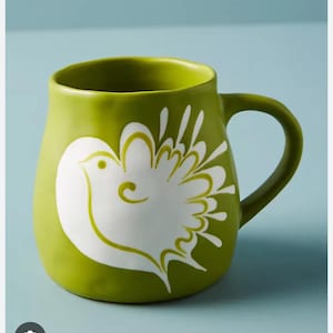 May include: A green ceramic mug with a white bird design. The mug has a rounded shape and a large handle. The mug is likely used for drinking beverages like coffee or tea.